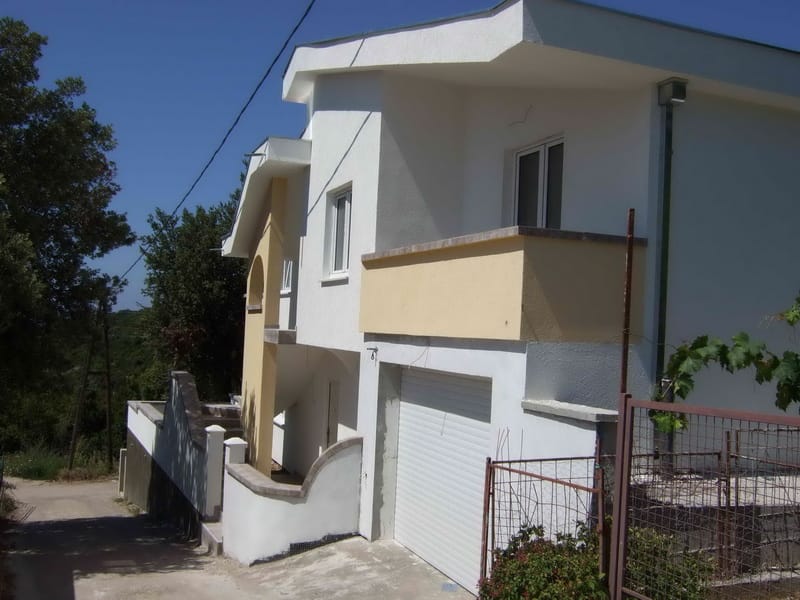 Two-story house with a garage and balcony in Uteha, Montenegro: building facade with white trim and adjacent road.