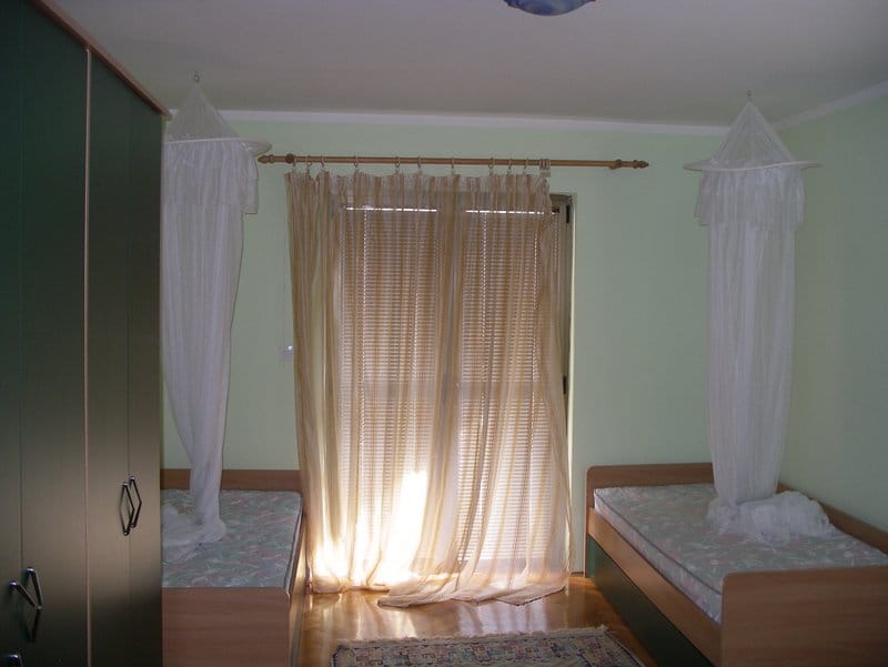 Bedroom in an apartment in Baošići, Montenegro: two canopy beds, a wardrobe, light walls, and access to a balcony with blinds.