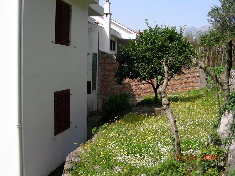 White house with red shutters and a green garden by the house in Dobrota, Montenegro. View of the yard and mountains.