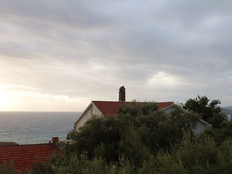 Sea view and a house with a red tile roof in Sveti Stefan, Montenegro, surrounded by greenery on a cloudy day.