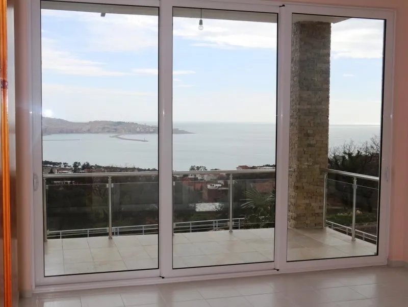 View from an interior room looking out through large glass sliding doors onto a tiled balcony with a stone pillar and sea view.