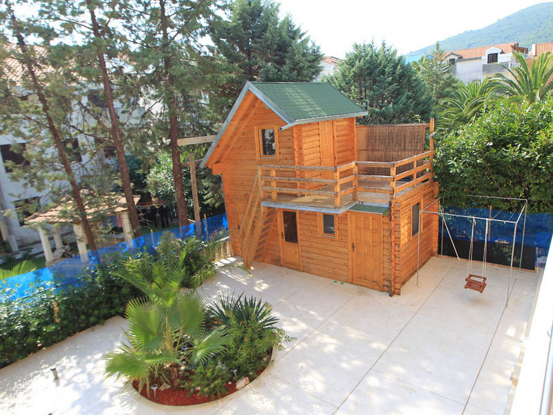 A two-story wooden cottage with a terrace and swings in the hotel courtyard in Budva, Montenegro, surrounded by greenery and palm trees.