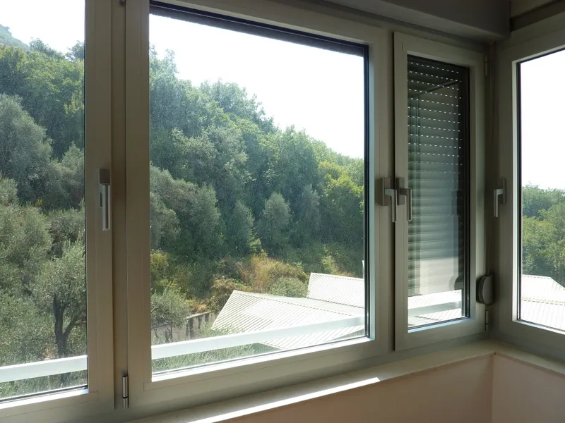 White window frames with handles and built-in blinds, overlooking a green hillside and rooftops.