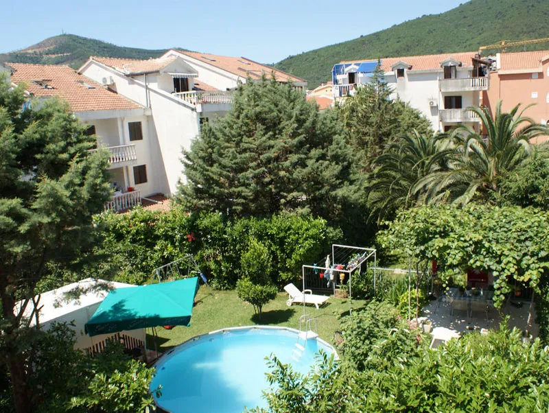 Overview of a round swimming pool in a garden surrounded by trees and residential buildings with red roofs near mountains.