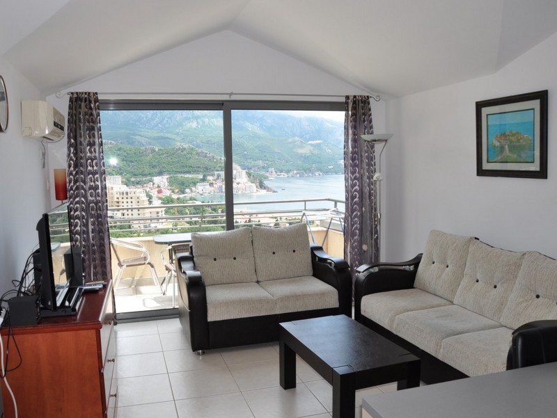 Living room in an apartment in Bečići, Montenegro: panoramic sea and mountain views, light-colored furniture, access to the terrace.