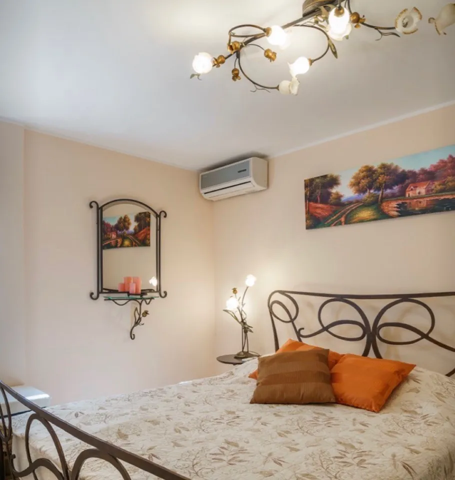 Bedroom featuring a metal bed frame, orange pillows, wall-mounted AC unit, and landscape artwork on beige walls.