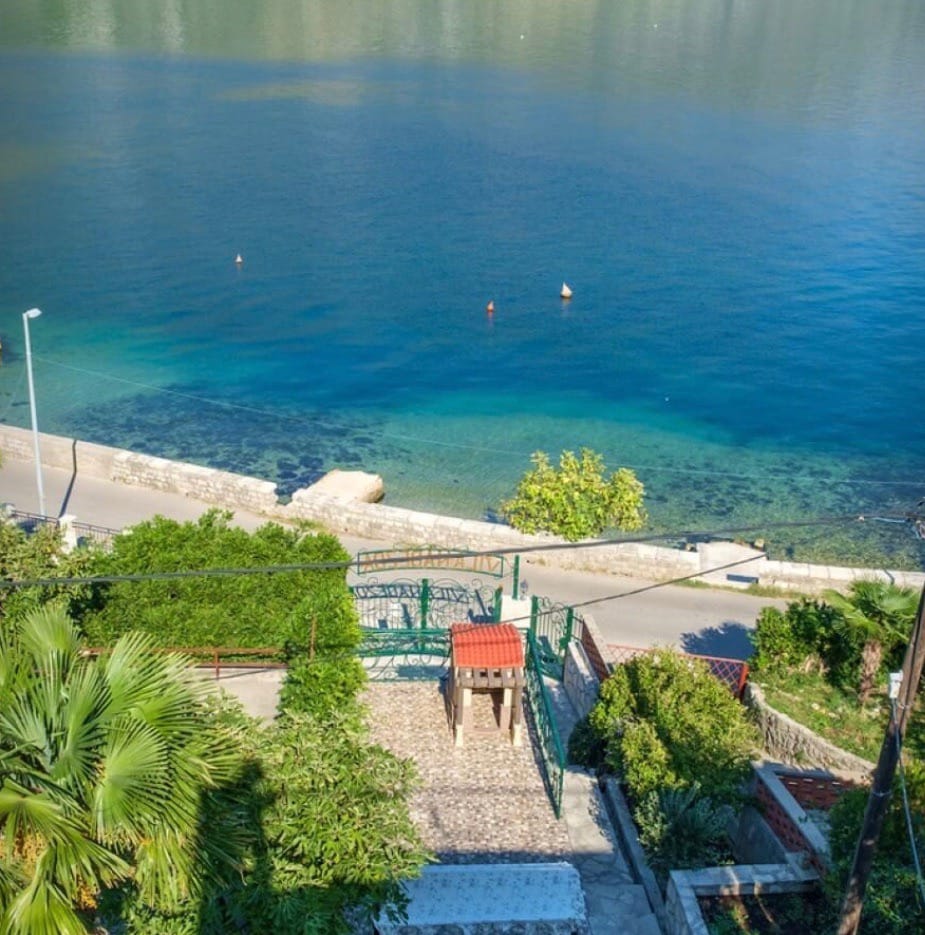 Sea and promenade view from the villa grounds in Stoliv, Montenegro: a green garden, stone path, and pier.