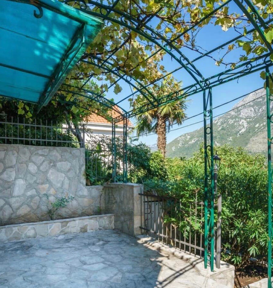 Terrace with a wrought-iron canopy and grapevine at a villa in Stoliv, Montenegro, overlooking the mountains and a green garden.