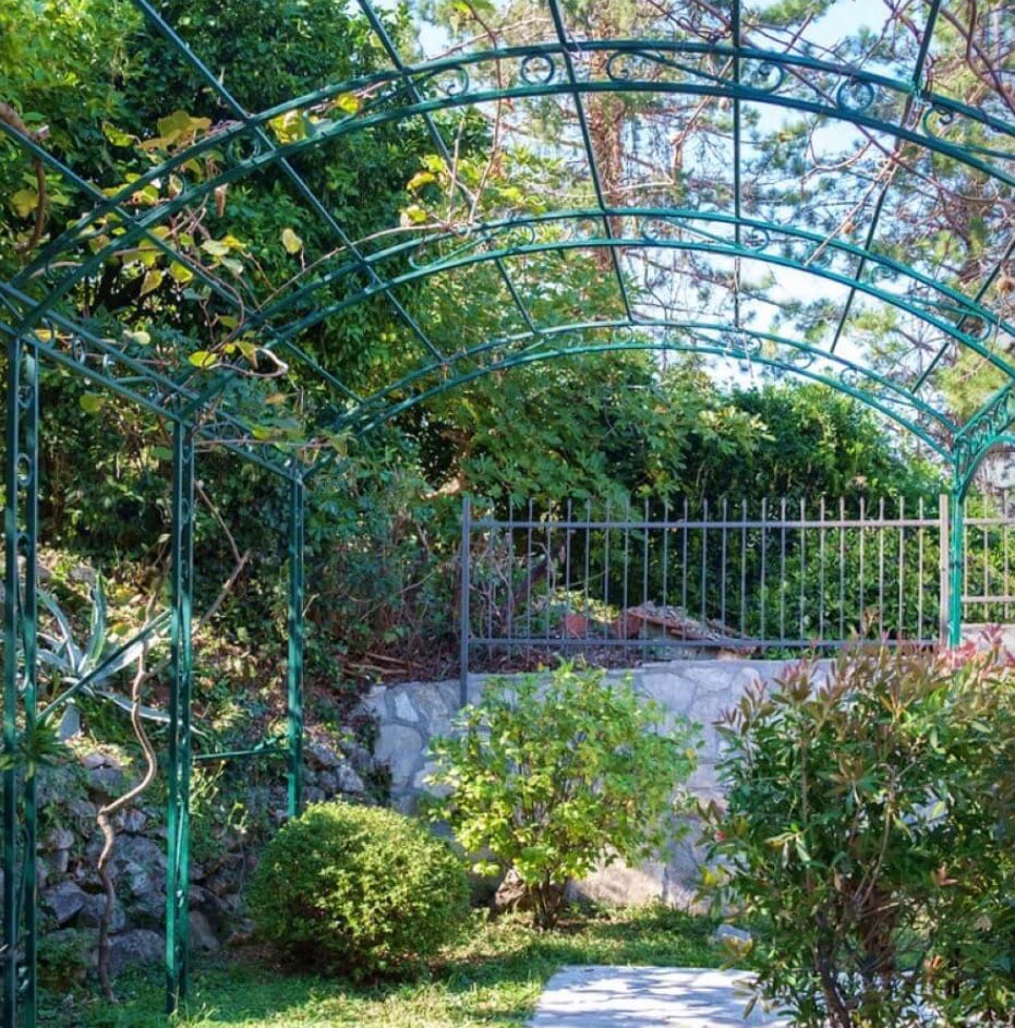Green wrought-iron arch in the garden of a villa in Stoliv, Montenegro, with a stone retaining wall and ornamental plants.