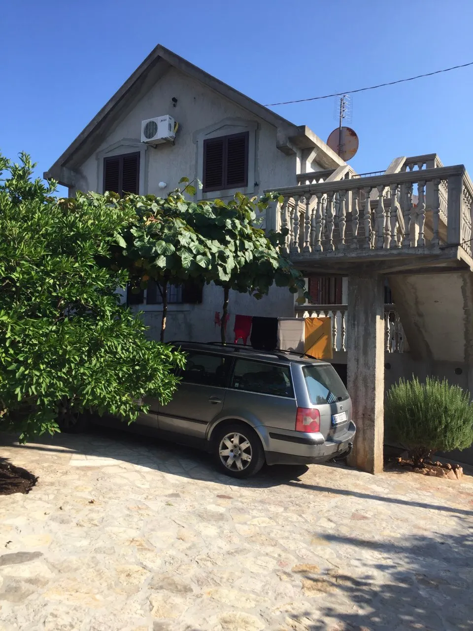 House exterior with stone driveway, silver station wagon, concrete balcony, green trees, and clear blue sky under bright sunlight.