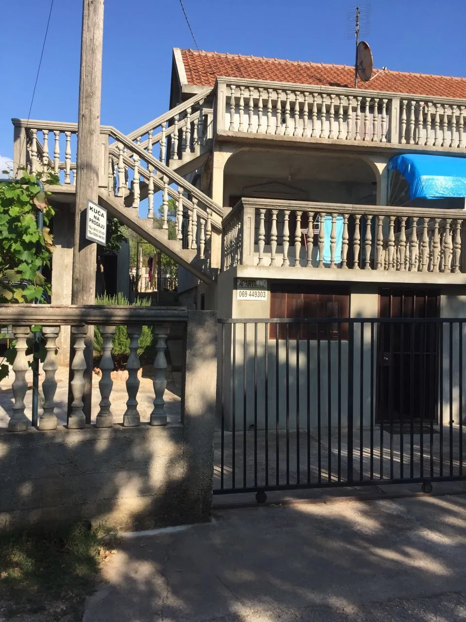Exterior view of a two-story house with balconies, concrete stairs, and a black metal gate under a blue sky.
