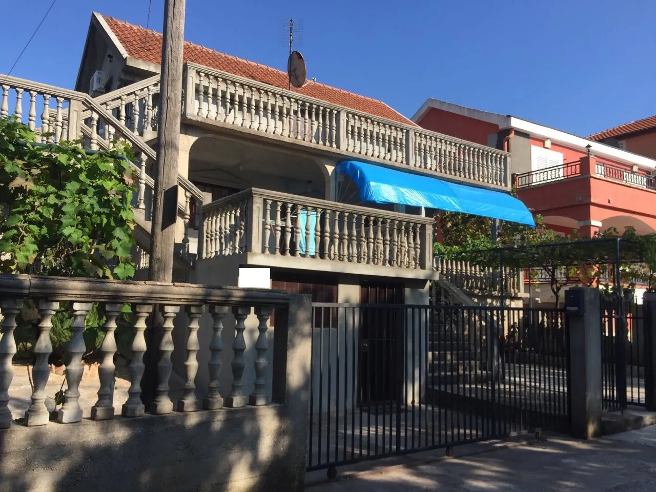 Exterior view of a two-story house featuring balconies with concrete balustrades, a blue tarp awning, and a black metal gate.