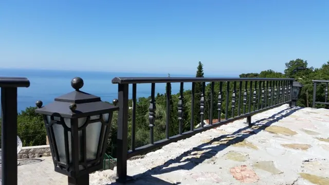 Stone balcony with black metal railing and lantern, overlooking the sea and trees under a clear blue sky.