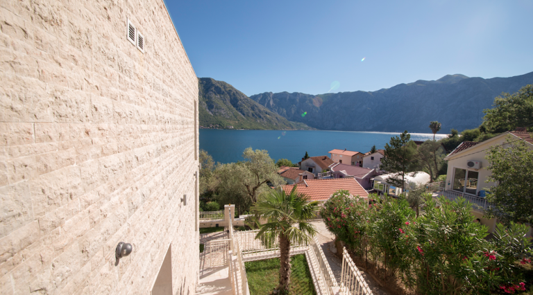View of the Bay of Kotor and mountains from the house terrace in Stoliv, Montenegro: stone facade and a well-kept garden with a palm tree.