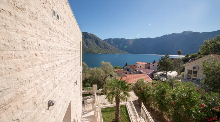 Stone house wall, garden with palm tree and flowering bushes, view of lake and mountains.