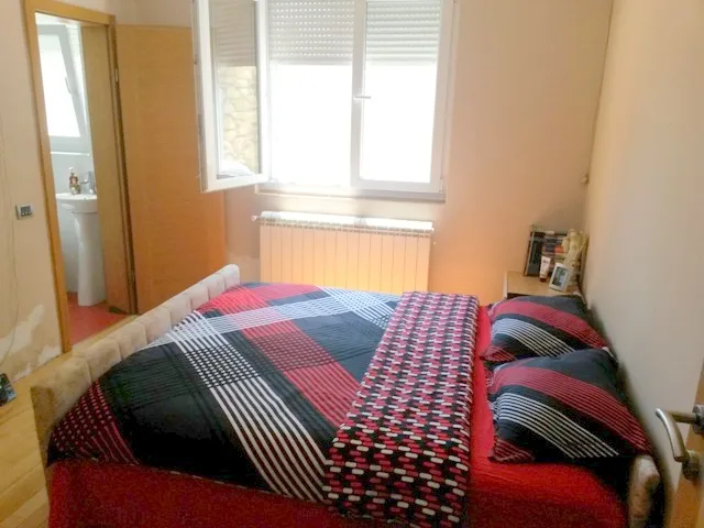 Bed with red and black plaid cover, window with radiator, and bathroom visible through door.