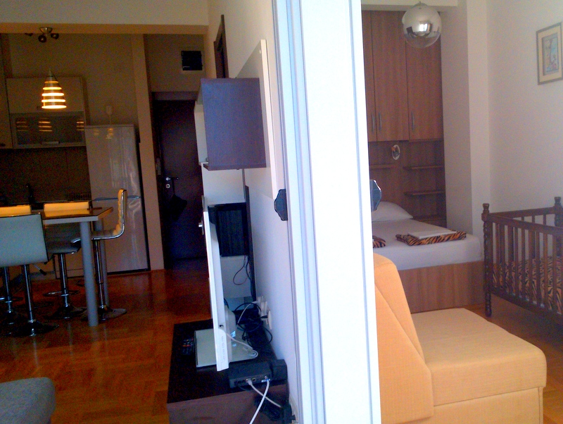 Apartment interior in Budva, Montenegro: a studio kitchen with a bar counter and a bedroom with a bed and a crib.