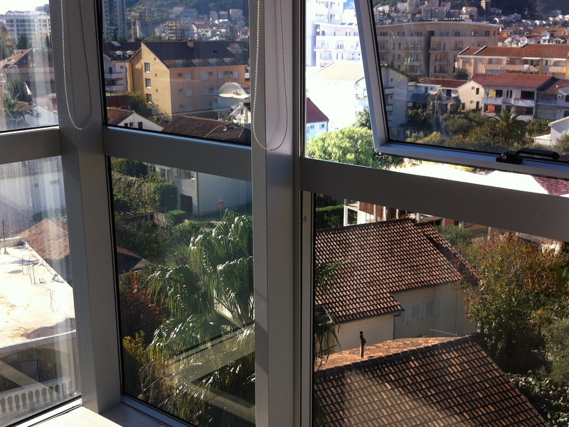 View from the apartment balcony in Budva, Montenegro: panoramic windows, tiled house roofs, and green palm trees against the city backdrop.