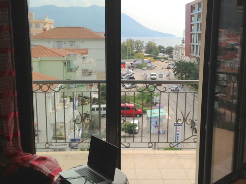 View from the apartment balcony in Budva, Montenegro: panoramic view of the city, mountains, and sea, wrought-iron railing, laptop on the table.