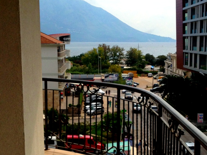 View from the apartment balcony in Budva, Montenegro: wrought-iron railing, city buildings, parking, and mountains against the sea.