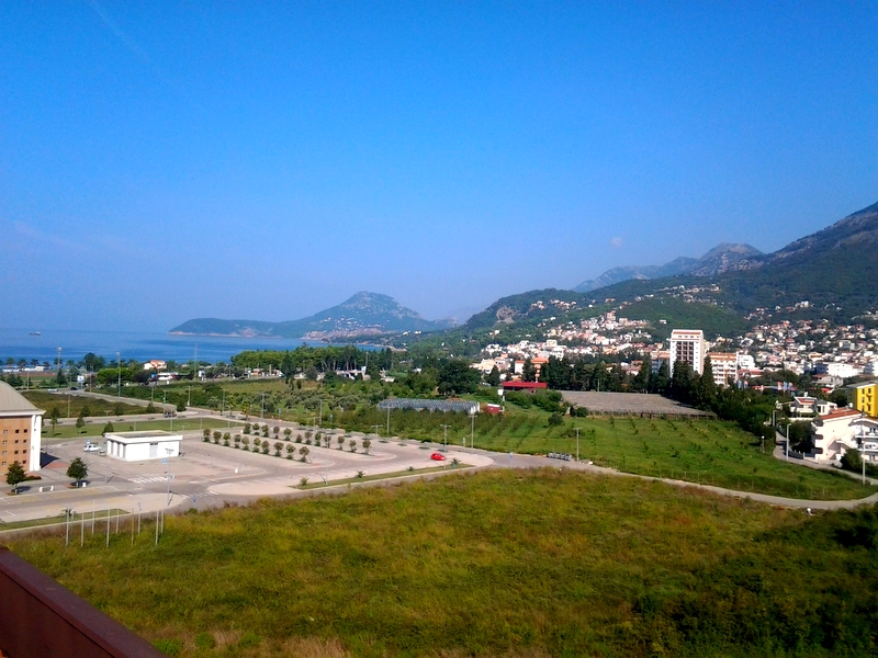 Panoramic view of the sea, mountains, and the city of Bar in Montenegro from the apartment window, with green fields and parking in the foreground.