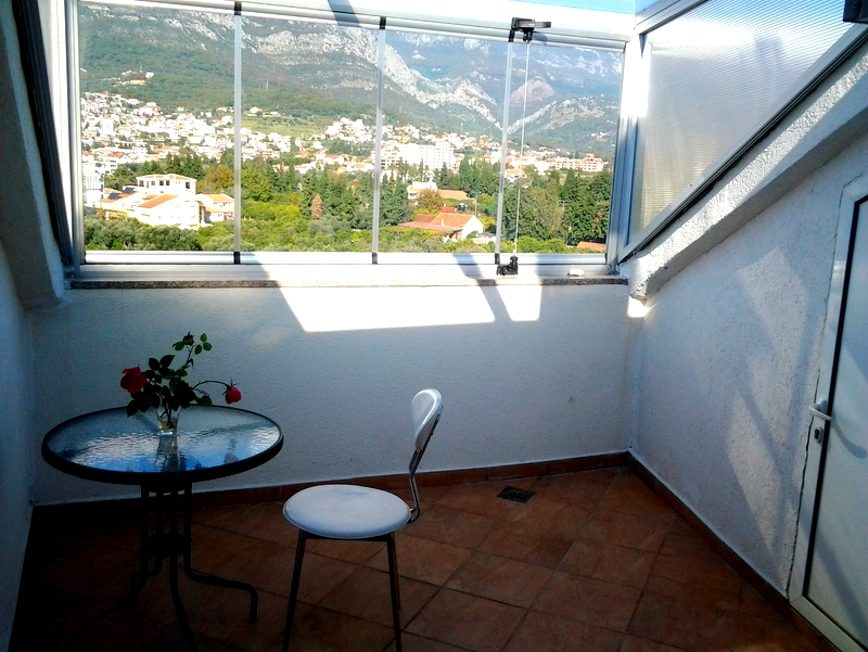 Balcony with panoramic views of the mountains and the city of Bar, Montenegro, with a glass table and chair in a bright apartment.
