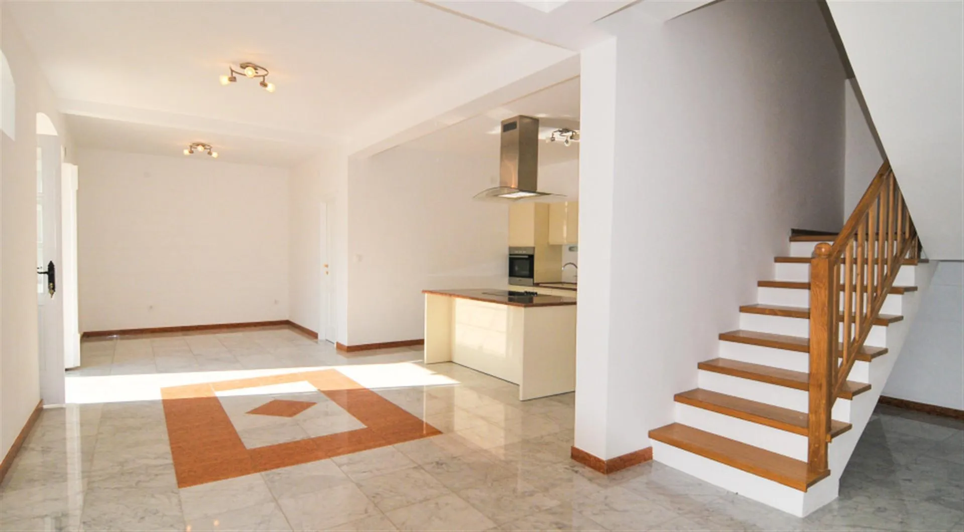 Spacious room with decorative floor tiles, a wooden staircase on the right, and an open kitchen area visible in the background.