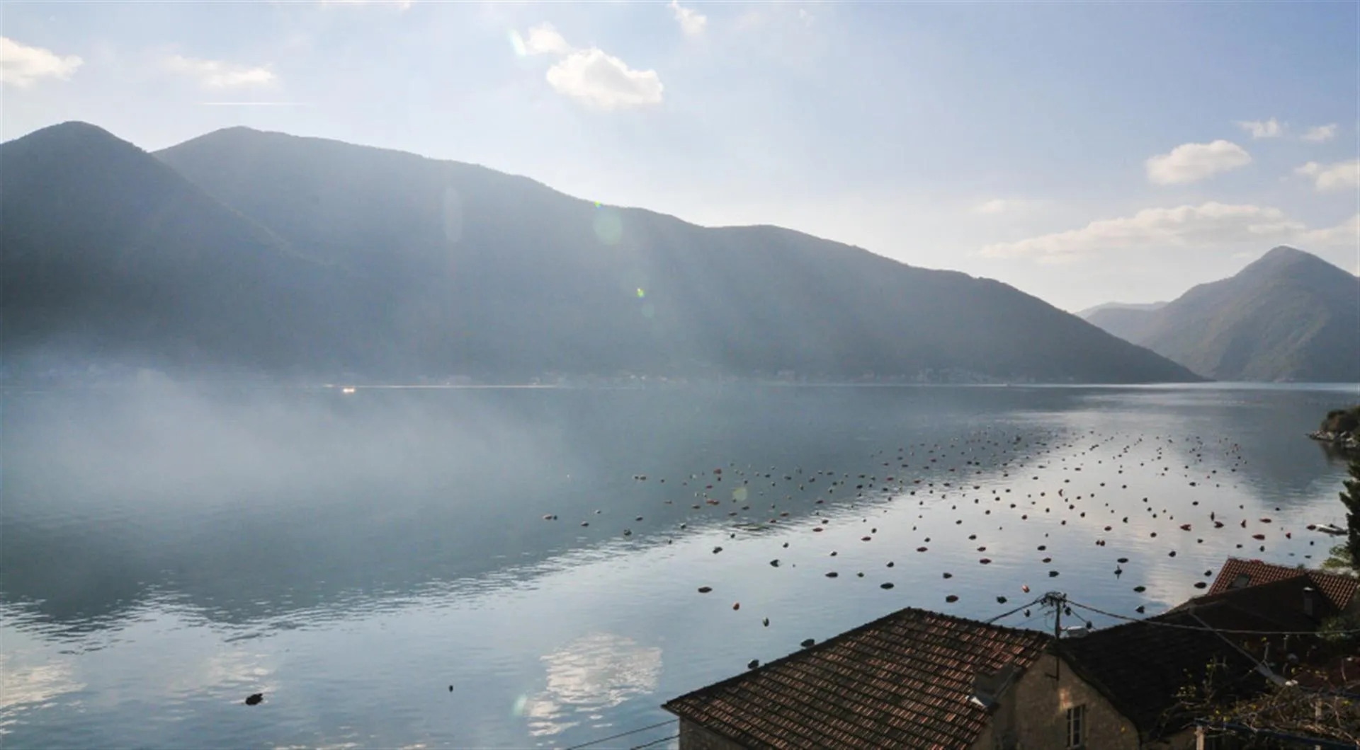Scenic view of a bay with mountains, floating buoys, and tiled rooftops in the foreground.