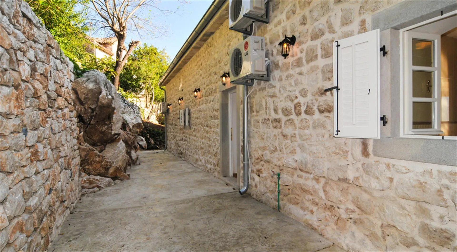 Stone house wall with AC units and lanterns along a narrow outdoor stone pathway under open sky.