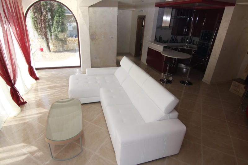 Living room with a white sofa and kitchen in a villa in Lastva, Montenegro: panoramic window, tiled floor, and stylish interior.