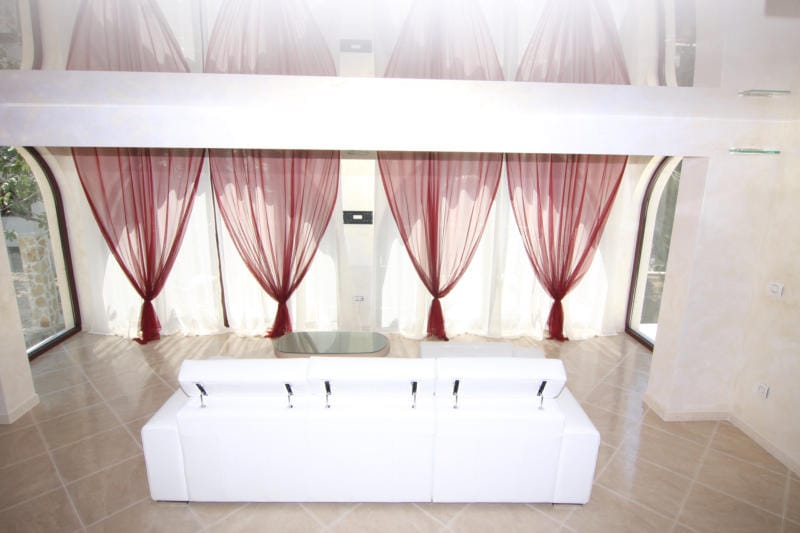 Villa living room in Lastva, Montenegro: white sofa, panoramic windows with red curtains, and a glossy ceiling.