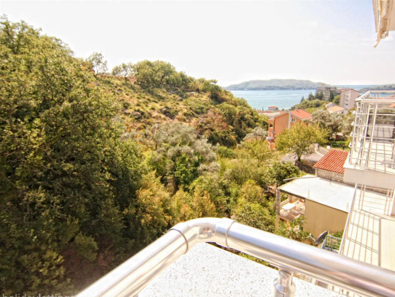 View from the apartment balcony in Bečići, Montenegro: a panorama of the sea, green hills, and residential buildings with terraces.
