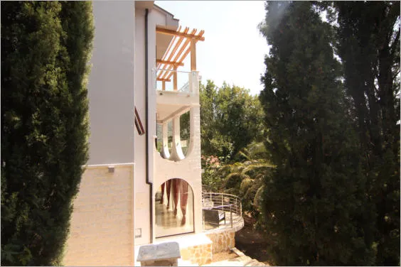 Exterior view of a house featuring a balcony, wooden pergola, and tall evergreen trees on both sides.