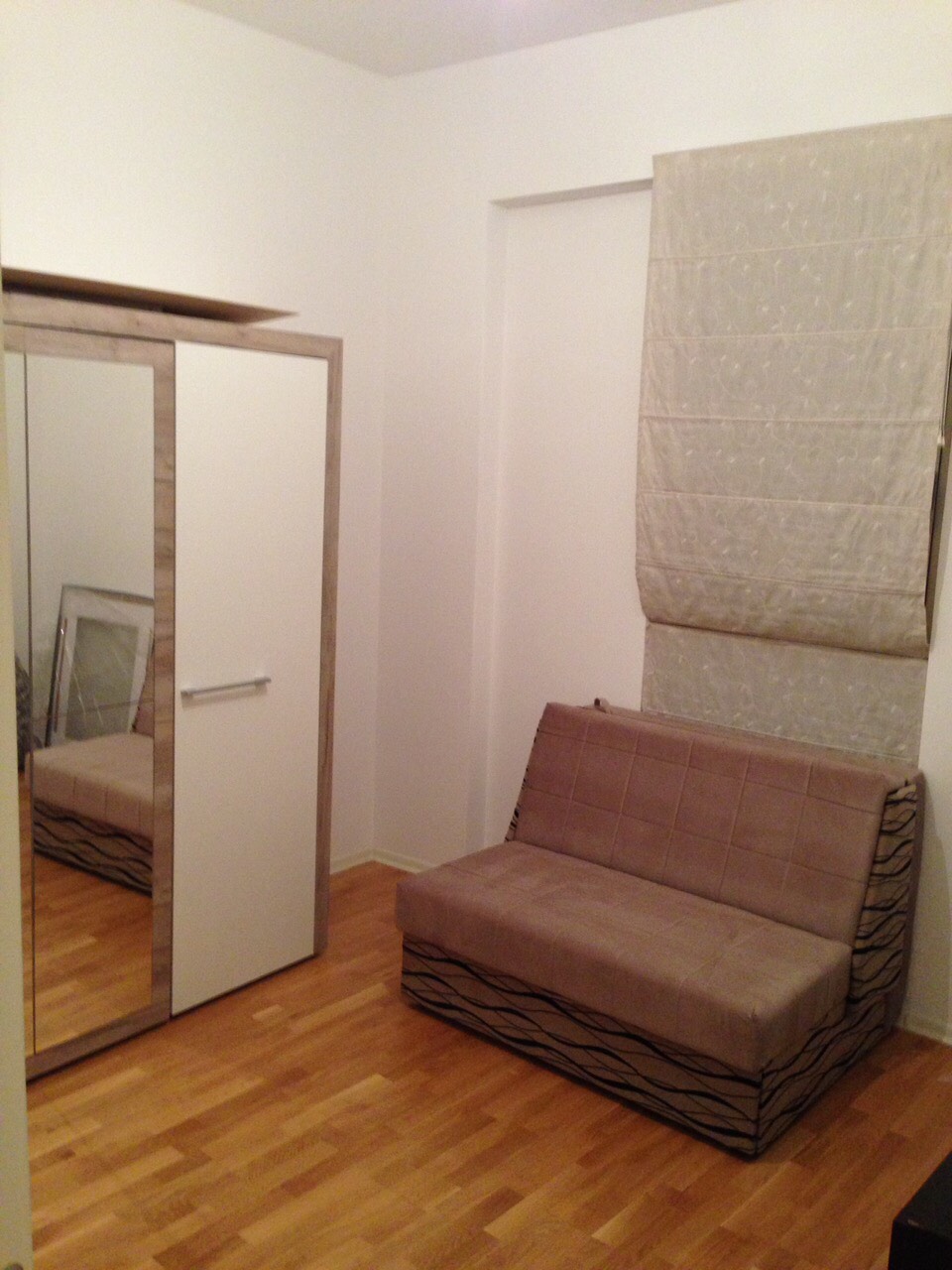 Bedroom in an apartment in Bečići, Montenegro: a light-colored mirrored wardrobe, a sofa bed, Roman shades, and parquet flooring.