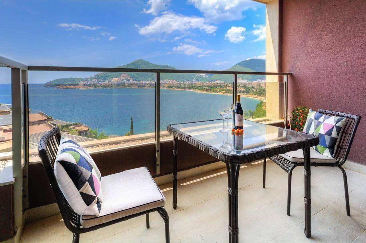 Balcony with sea and mountain views in an apartment, Becici, Montenegro: wicker chairs, a glass table, and panoramic glazing.