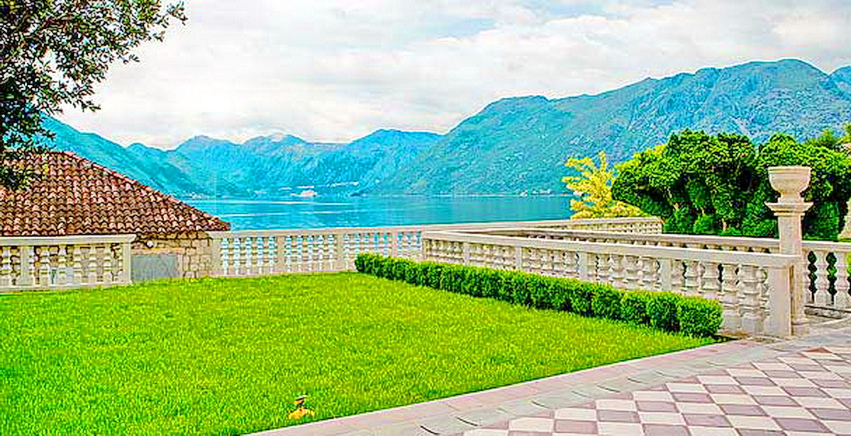 View of Kotor Bay from the villa terrace in Dobrota, Montenegro: a well-kept lawn, stone balustrade, and mountains.