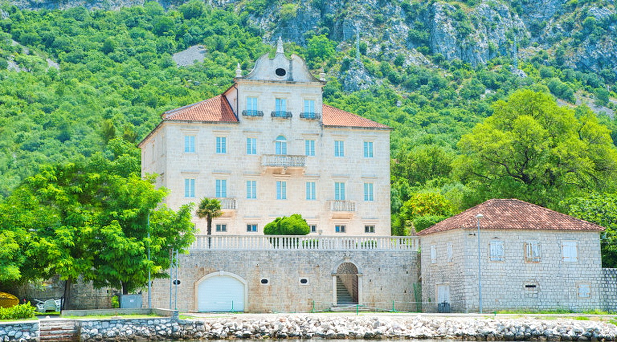 Historic stone villa in Dobrota, Montenegro: building facade with a tiled roof against a backdrop of mountains and greenery.