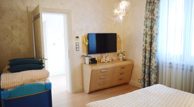 Cozy bedroom in a villa in Tivat, Montenegro: light furniture, TV, designer chandelier, and a view of the adjacent room.
