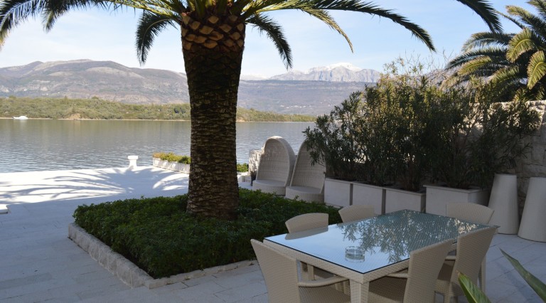 Villa terrace in Tivat, Montenegro: dining area, palm trees, sea view, and mountains with snow-capped peaks.