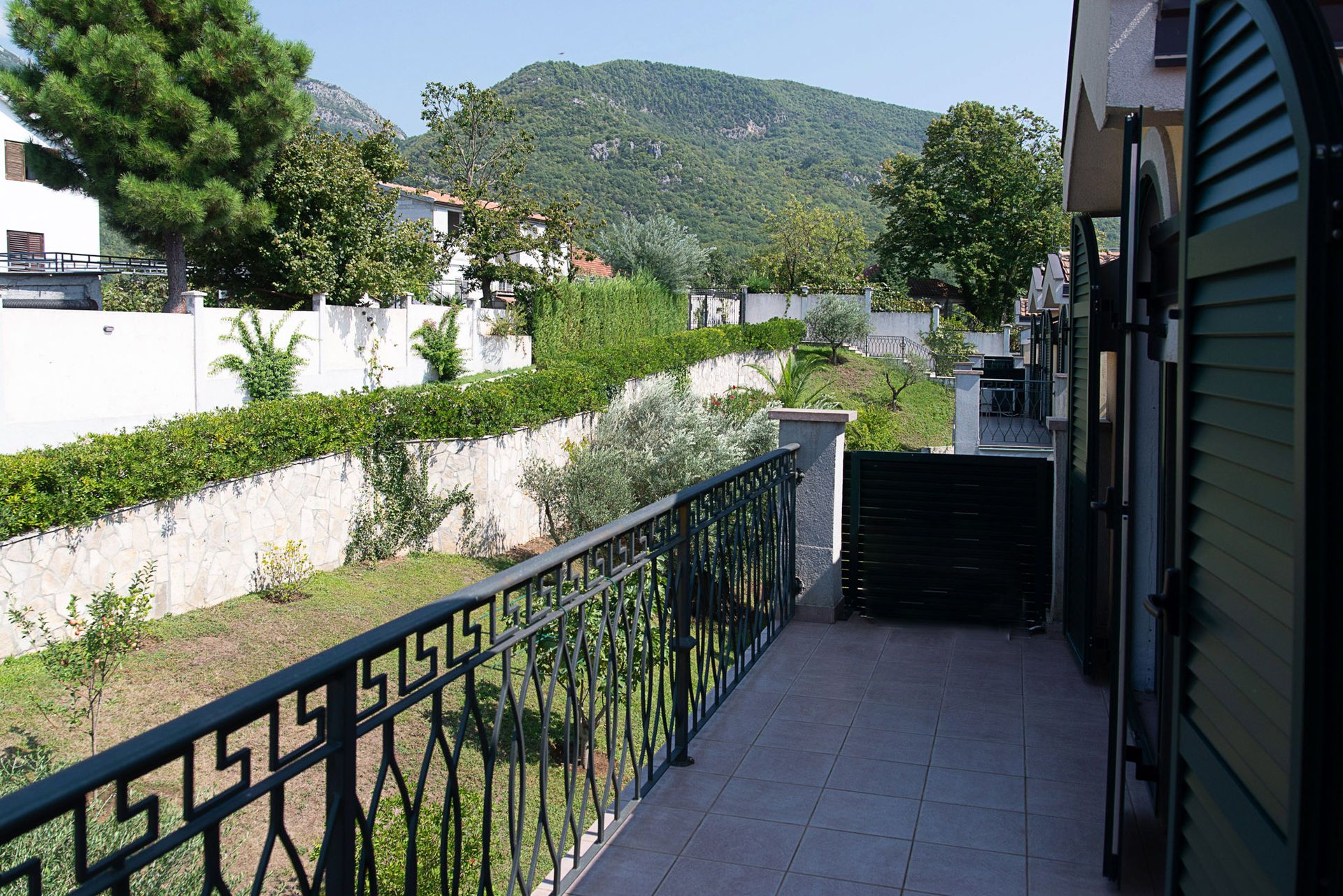 Townhouse balcony in Lastva, Montenegro, with a view of green mountains, a garden, and wrought-iron railing.