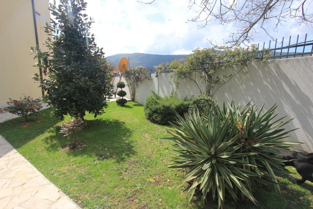 A well-kept green garden with ornamental plants by a townhouse in Lastva, Montenegro, with mountain views.