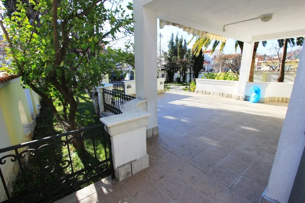 Covered terrace with tiled floor, white columns, and black wrought iron railing overlooking a garden area.