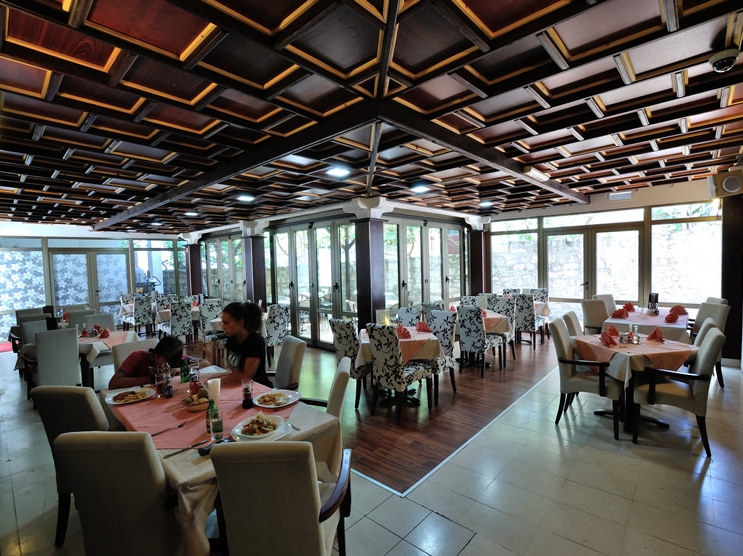 Hotel restaurant interior in Petrovac, Montenegro: coffered wooden ceiling, panoramic windows, and upholstered chairs.