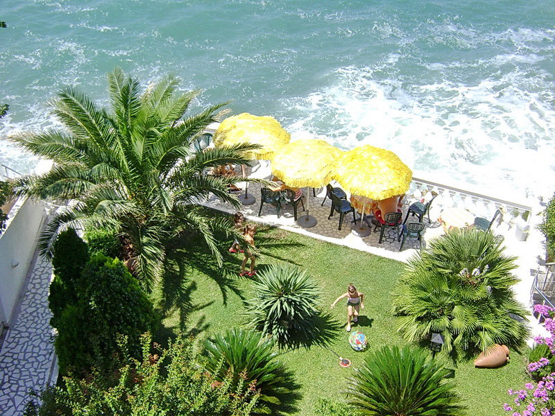 Top view of the hotel’s green garden in Sutomore, Montenegro, with palm trees, yellow umbrellas by the sea, and a lounge area on the lawn.