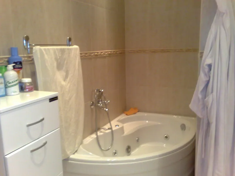 Corner jacuzzi tub, white cabinet with toiletries, towel on rack, beige tiled walls visible.