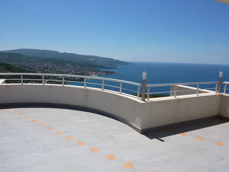 Wide tiled balcony with metal railing overlooking the sea and a mountainous coastline under blue sky.