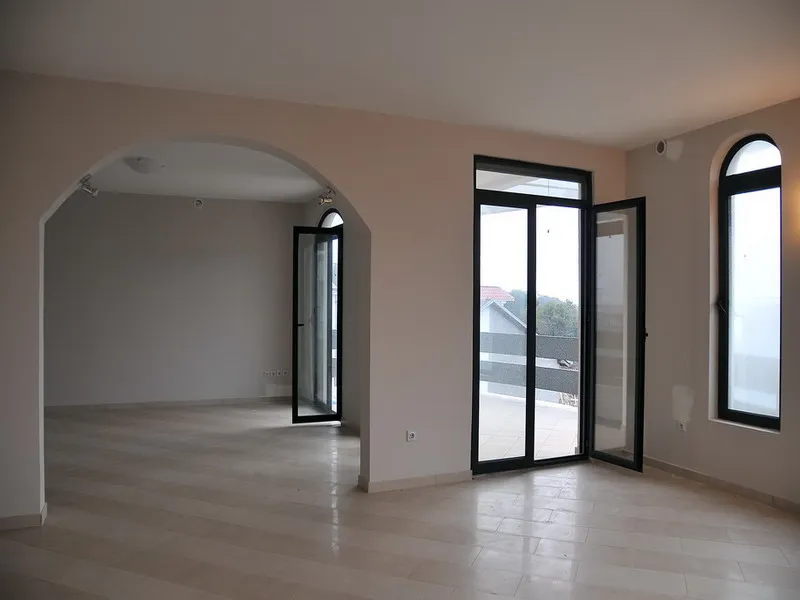 Empty room with an arched doorway, light tiled floor, and large black-framed windows opening to a balcony.