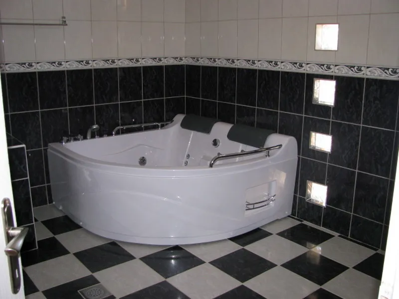 Large corner jacuzzi bathtub on black and white checkered floor with dark tiled walls and built-in lights.