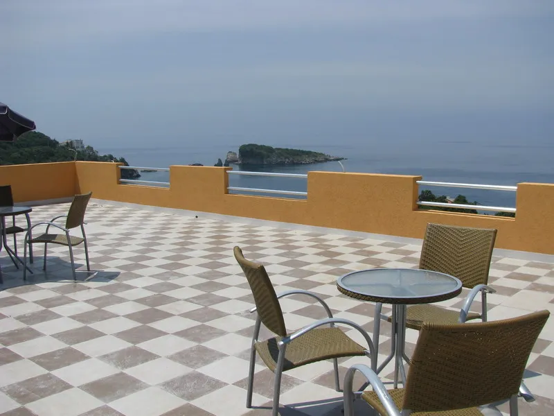 Terrace with checkered tiles, wicker furniture, and a view of the sea and island.