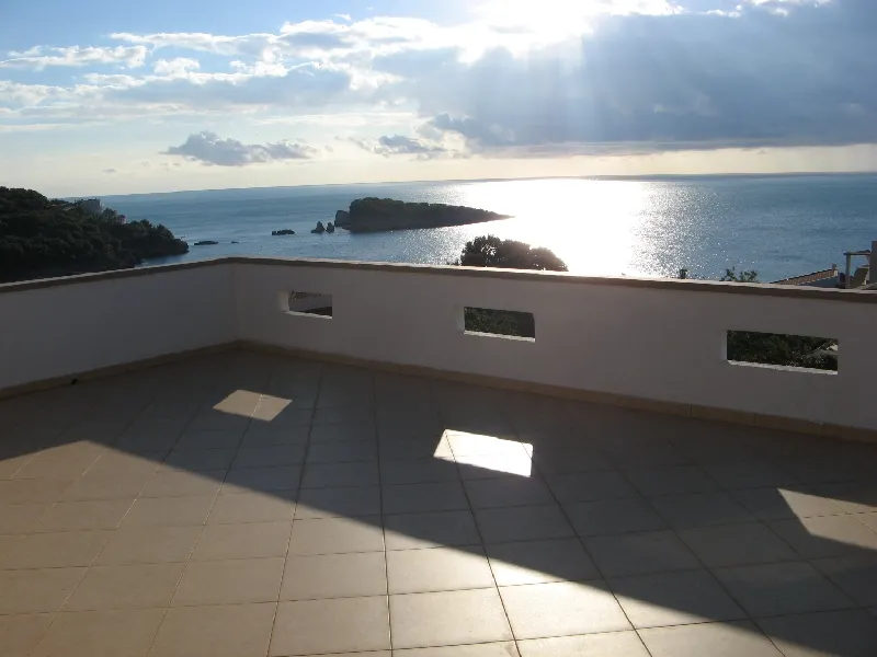 Open terrace with white walls and tiled floor overlooking the sea and island under sunlight.